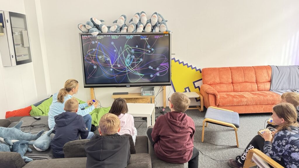 Kinder und Jugendliche sitzen vor einem großen Bildschirm, auf dem das Spiel "Newtonwars" gezeigt wird.