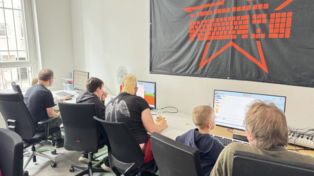 Jugendliche sitzen mit Helfenden vor Monitoren, die das Internetangebot "Programmieren mit der Maus" zeigen.